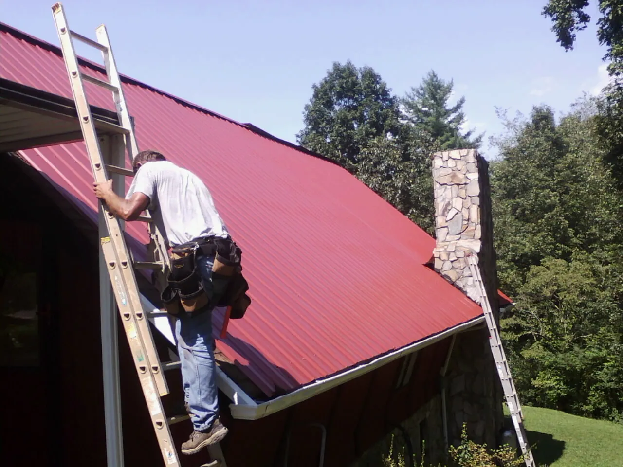 Roofer on a ladder beside a red metal roof and stone chimney — Soffit & Fascia Repair by Roofing Brothers in Bingen